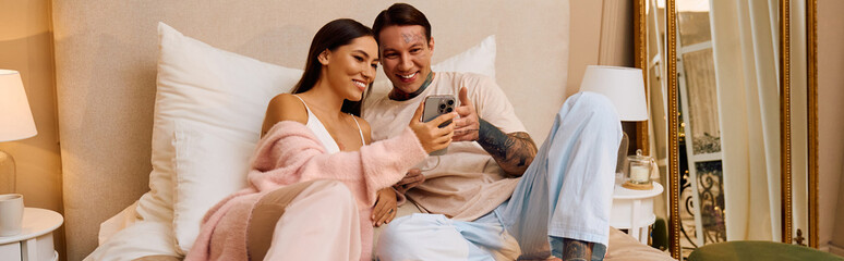 Young couple cuddles together on a bed, sharing laughs while looking at a phone screen.