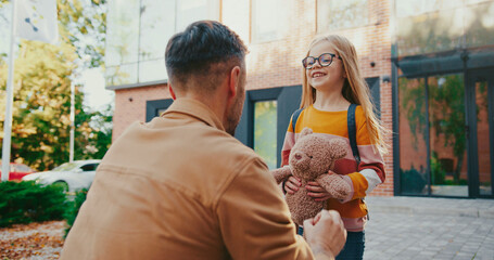 Camera view Caucasian dad expecting his lovely daughter after school lessons. Pretty girl with teddy bear in her hands running to father. Child sharing good feelings and telling news during day.
