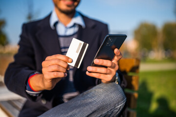 Close up photo of businessman using mobile phone and credit card while sitting on the bench in the park. Focus on phone in hand. 