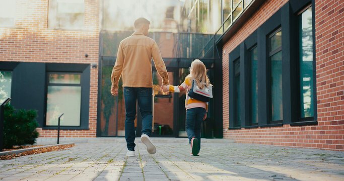 Warm moment of parent holding hand of his small child. Kid jumping while walking together with father towards school entrance. Daughter happy about starting studying in new location.