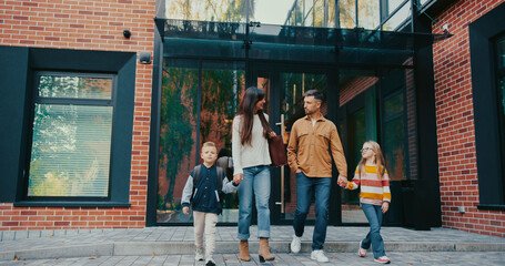 Front camera view of Caucasian family walking together away from school back home. Wife holding hands with son and dad holding hands with daughter. Kids happy about spending time with parents.