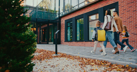 Camera view from behind small park area. Kids with backpacks running to school entrance. Parents waving their hands to children. Saying goodbye. Childhood concept. Yellow leaves on ground.