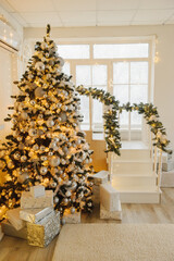 A tall Christmas tree with gold and silver ornaments is the centerpiece of a cozy living room. The tree is surrounded by presents and a garland adorns the nearby staircase. The room has a festive and 