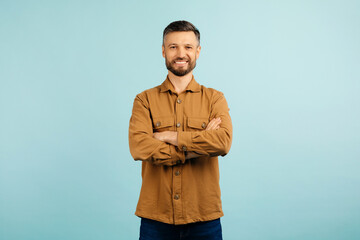 A cheerful caucasian middle aged man stands with arms confidently crossed, wearing a casual brown shirt, against a solid blue background. High quality photo