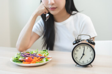 Intermittent fasting, close up clock asian young woman, girl diet, waiting time to eat ketogenic low carb, green vegetable salad on plate. Eat food healthy first meal in brunch, lunch on table at home