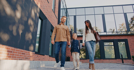 Happy family walking in front of brick building with modern architecture. Beautiful mother with long hair holding her son hand. Father on other side looking cheerful. Boy in middle carrying backpack