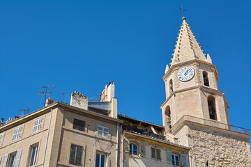 The clock tower of Notre-Dame-des-Accoules in Marseille, France, with its distinctive spire and clock face, set against a clear blue sky and surrounded by traditional buildings.