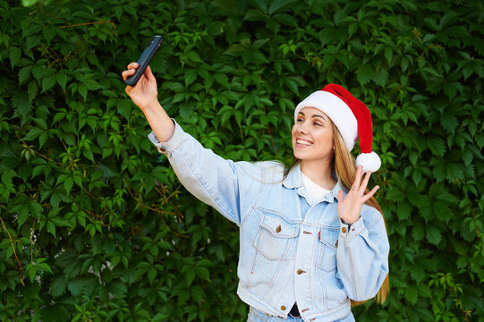 Caucasian young woman wearing santa claus hat with smartphone talking on video call on green background. Woman with phone in Santa hat on green background. Background of leaves. Christmas. New Year.