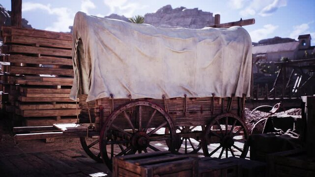 A covered wooden wagon rests in an old west settlement. It is draped with a cloth and surrounded by wooden structures under a bright sky, evoking a historical atmosphere.