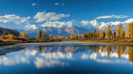 Naklejka premium Stunning panoramic landscape showcasing serene lake reflection of majestic snow-capped mountains against a vibrant blue sky with scattered clouds and golden foliage