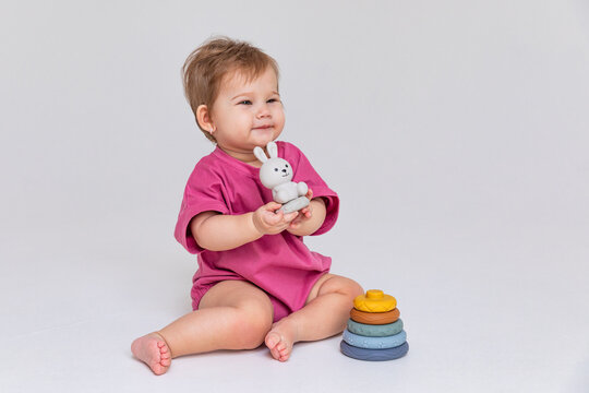 Happy smiling little baby girl, up to one year old, sits on the floor and plays with a multi-colored pyramid sorter on a white background. Development of fine motor skills. Early education. Copyspace
