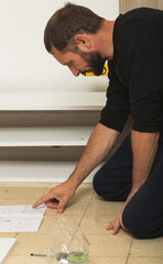 Man kneels on the floor, intently examining assembly instructions for a furniture piece. His focused expression and precise hand gesture suggest careful attention to detail, in a home improvement task