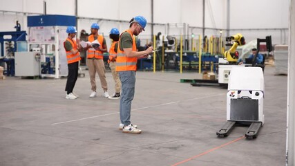 Multiracial team of industrial engineers testing an automated guided vehicle - agv - and robotic arm in a large modern factory, writing notes and discussing production process optimization