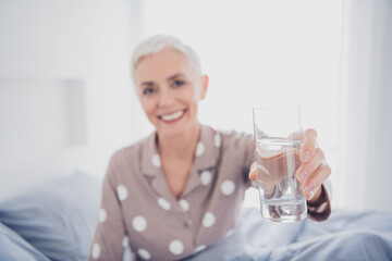 Beautiful mature woman waking up in the morning holding a glass of water indoors, enjoying comfort and relaxation in her cozy bedroom