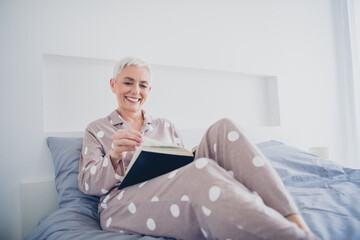 Elderly woman in pajamas enjoying reading in bed, capturing the essence of leisure and comfort in a bright, tranquil bedroom setting