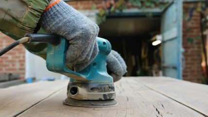 A man in camouflage work clothes and gray gloves works textured wood with cracks with a blue hand sander against a brick garage with green vegetation on it and an open blue door