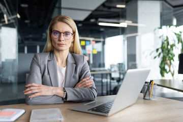 Mature businesswoman seated in modern office, wearing glasses and a gray suit, demonstrating confidence and leadership. Laptop and tablet on desk, symbolizing professionalism and success.