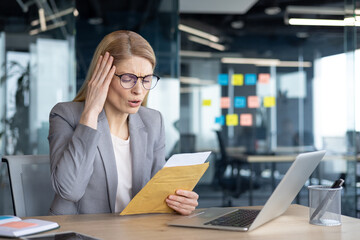 Obraz premium Mature woman businesswoman reading bad news letter looking worried. Seated at desk with laptop, showing stress in office. Concept of business challenges and unexpected setbacks.