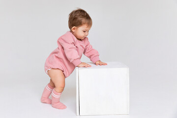 The child baby girl in pink bodysuit takes her first steps and stands near a large white cube on a...