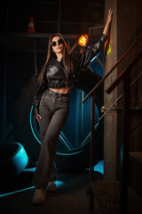 A beautiful woman in jeans and a black leather jacket in neon light posing on metal concstructions background.