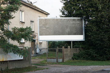 Billboard on private property near a concrete house. Empty banner for roadside advertising. Blank copy space for promotional design. Outdoor advertisement background.
