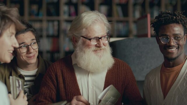 Elderly author with white beard standing in library, surrounded by group of enthusiastic fans holding copies of his book, putting signature on title pages and answering questions from readers