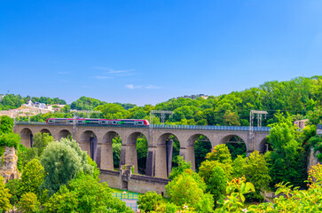 Fototapeta premium Train moving on viaduct bridge Pulvermuhl Viaduct Biisser Breck railway stone arch across Alzette valley in Luxembourg City historical centre, park with green trees and blue sky background