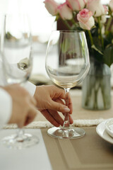 A close-up of a hand holding an empty wine glass with a bouquet of pink roses in the background on a dining table setting