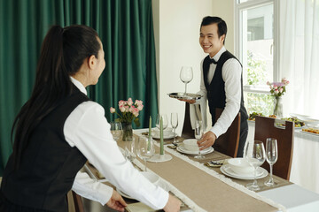 Waitstaff setting up dinner service in an elegant dining area, arranging glassware and utensils on a stylishly adorned table with flowers and a cheerful ambiance