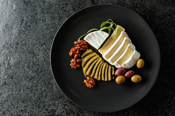 Camembert cream cheese with pesto, olives, rosemary, and walnuts on a black plate.