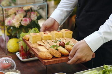 Person holding a wooden tray filled with assorted appetizers, surrounded by a well-decorated table and flowers. The scene exudes an elegant and refined ambiance