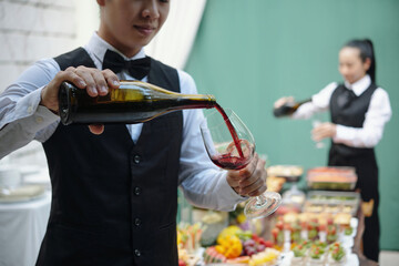 Waiter pouring red wine into glass at elegant outdoor event with food and drinks. Background features another server preparing refreshments
