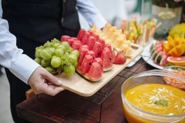 Person arranging colorful slices of watermelon, pineapple, and grapes on wooden platter set on...