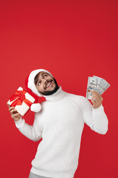Funny hipster man wears santa claus hat holding gift box and cash money over red background. Looking up. Copy space.
