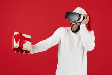 Shocked hipster man wears santa claus hat holding gift box and wearing VR headset goggles over red background.