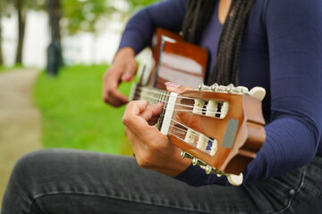 Woman with Brown Skin and Long Black Hair with Dreadlocks Sitting on Bench Playing Guitar in Green Park. Medium Shot