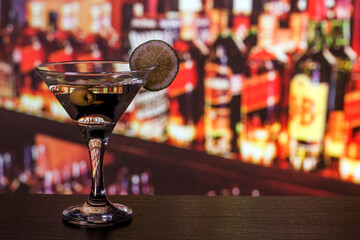Martini glass with olive and lime on the bar counter. In the background there is a bar with bottles of alcohol