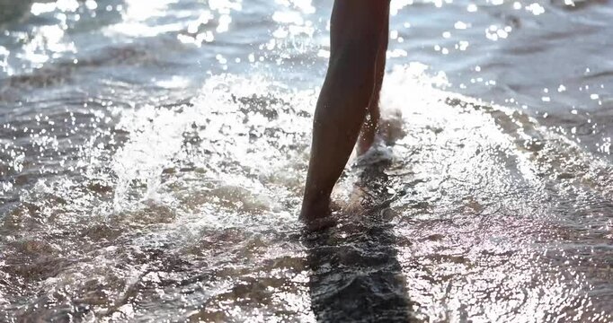 Legs of woman walking along edge of beach washed by sea waves slow motion. Sun reflected in reams of water creating tantalizing shine