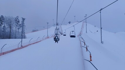 The snow-covered slopes with lampposts for skiers and snowboarders at the ski center are a mesh fence for safety and surrounded by a forest. Skiers go up the chairlift. Overcast and dense cloudiness