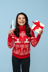 Surprised young brunette woman dressed in Christmas sweater opening gift box over blue background.