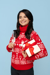 Smiling young brunette woman dressed in Christmas sweater opening gift box over blue background.