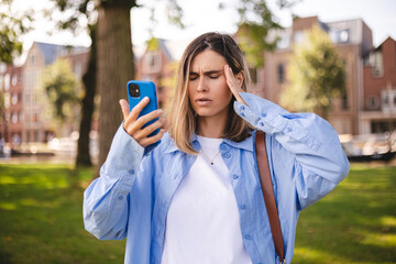 Blonde short hair woman wear blue shirt, hold mobile phone touch her head and rolling her eyes,...