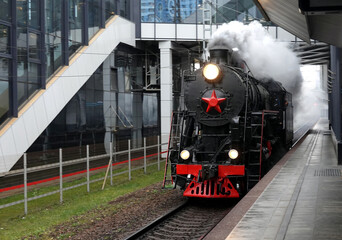 Industrial landscape with old black steam locomotive with red star on front arriving to the railroad station in cloudy day perspective view © DyMaxFoto