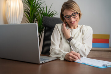 Cheerful mature businesswoman sitting at her workplace. Happy mature businesswoman working at her desk in the office. Smiling mature businesswoman focused on her tasks at work.
