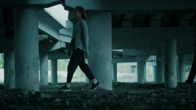 Full lowkey shot of young lady walking under bridge and suspicious masked man pursuing her and looking around, human trafficking