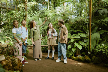 Wide shot of young female botanical garden worker demonstrating various plants growing in greenhouse to group of teen students