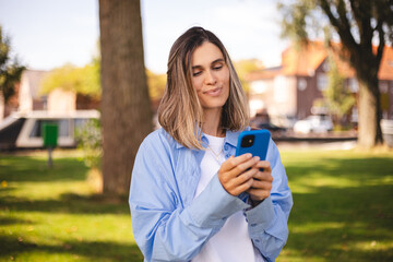 Attractive blonde short hair woman wear blue shirt hold mobile phone and look happy and smiling. Amazing enjoy woman walk in the park. Girl scrolling, messaging, write sms, read news, think answer.