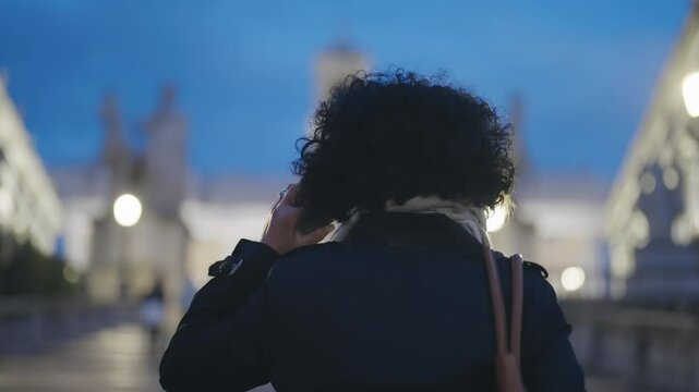 A business woman communicates talking on phone while walking at the Cordonata Capitolina with defocused Campidoglio at background in Rome during twilight, capturing the essence of work and travel.