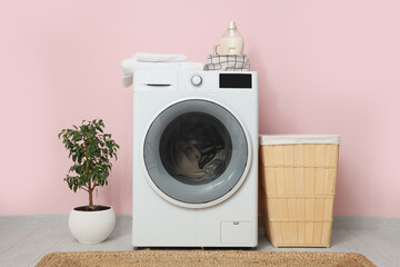 Modern washing machine with laundry, basket and houseplant near pink wall