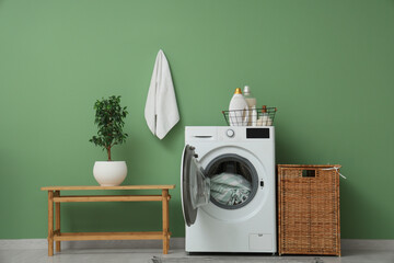 Modern washing machine with bench, detergents and basket near green wall